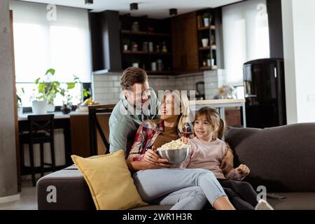 A family of three is comfortably nestled on a couch, their faces reflecting excitement and attentiveness as they share a bowl of popcorn during a susp Stock Photo