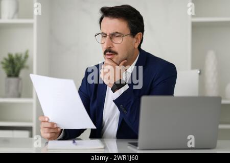 Serious businessman in a blue suit critically examining a document while sitting at his desk with a laptop Stock Photo