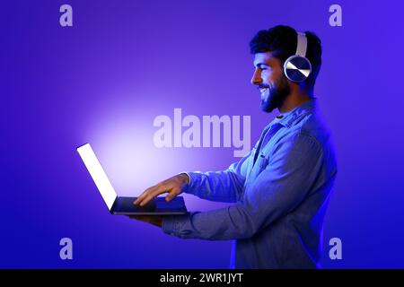 Bearded guy with earphones stands typing on laptop, neon background Stock Photo