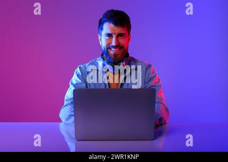 programmer guy sitting with laptop at desk in neon studio Stock Photo