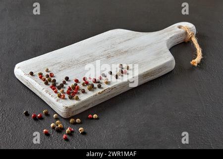 Wooden white cutting board with scattered colorful peppercorns on a black concrete countertop Stock Photo