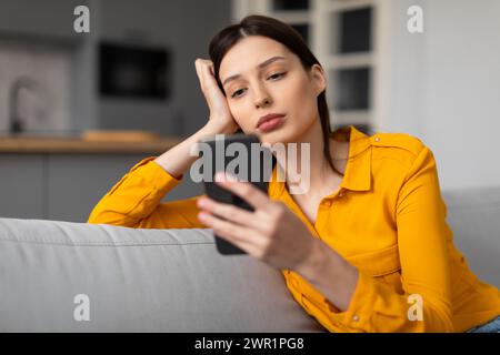Pensive woman with phone looking sad and thoughtful on sofa Stock Photo