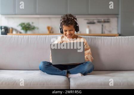 Happy black preteen boy wearing headphones using laptop at home Stock Photo