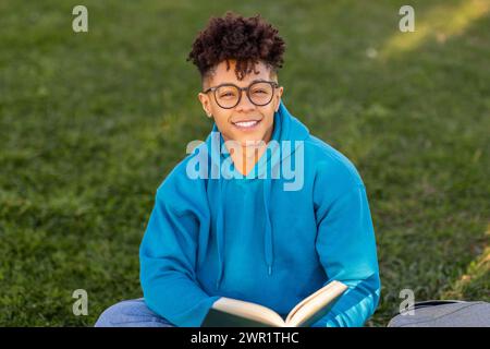 Brazilian student guy wearing glasses happily holding book in park Stock Photo
