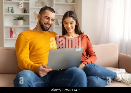 Technologies For Leisure. Happy Young Couple Using Laptop Together At Home Stock Photo
