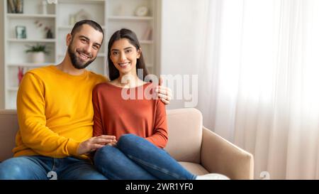 Loving man and woman sitting closely on sofa and smiling at camera Stock Photo
