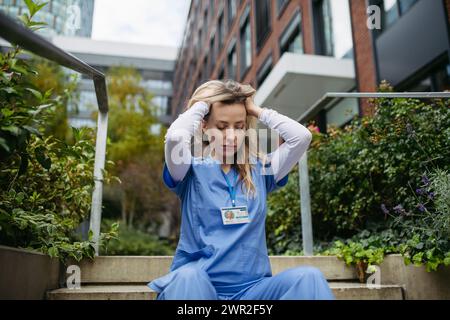 Female doctor feeling overwhelmed at work, sitting on stairs. Healthcare workers with stressful job, feeling exhausted. Burnout syndrome for doctors Stock Photo