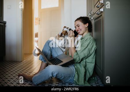 Young student e-learning on laptop at home, cute small dog wants to play. Businesswoman working remotely, sitting at kitchen floor, phone calling Stock Photo