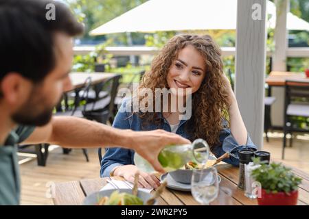 Young couple at date in restaurant, sitting on restaurant terrace. Boyfriend pouring water in girlfriends glass, taking care of her. Stock Photo