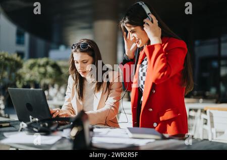 Two focused women engaged in work at an outdoor table, one using a laptop and the other on a call with headphones. Stock Photo