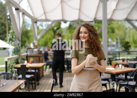 Portrait of a beautiful waitress writing down an order in paper notebook with pen. Server standing on restaurant terrace in an apron. Stock Photo