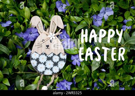 Greeting card with text Happy Easter in English. Easter decor. Decorative wooden Easter bunny toy on background of Vinca blooming with lilac flowers. Stock Photo