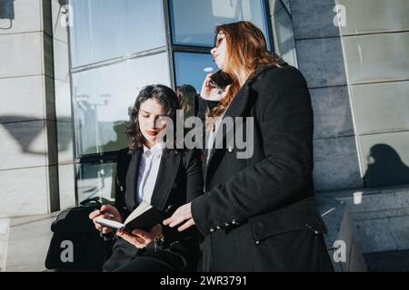 Two motivated young female entrepreneurs collaborate outside an office building, one on a phone call and the other taking notes. Stock Photo