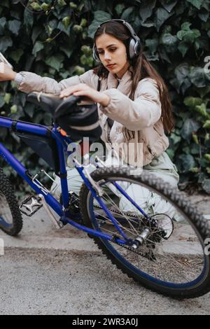 Focused woman with headphones attaching helmet to bicycle in urban setting. Stock Photo