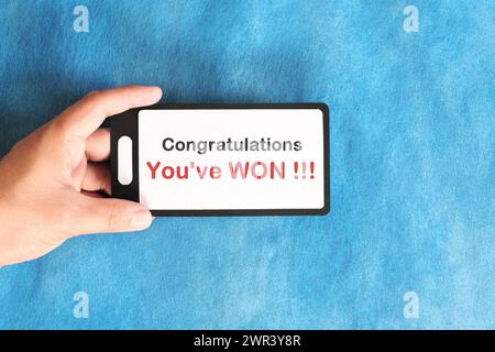 Winning text scam concept. Hand holding mobile phone with congratulatory message in blue background. Stock Photo