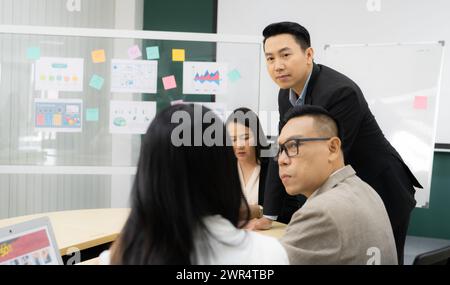 A group of people are gathered around a table with a whiteboard behind them. They are discussing something important, and one of the men is wearing Stock Photo