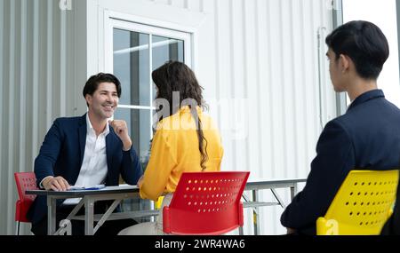 Three people are sitting at a table, with a man in a suit and tie smiling at the other two Stock Photo