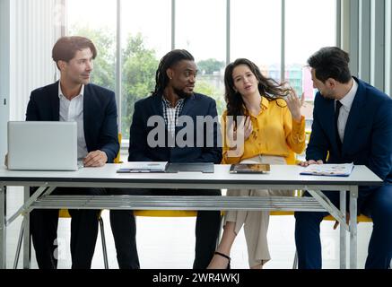 A group of people are sitting around a table in a business setting. One woman is talking to the group while the others listen. The atmosphere is profe Stock Photo