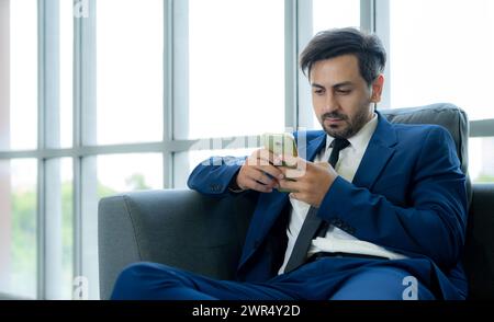 A man in a suit is sitting on a couch and looking at his cell phone. He is focused on the screen, possibly checking his messages or browsing the inter Stock Photo