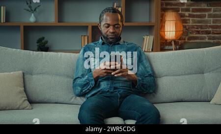 Alone African American man guy male sitting at hone couch sofa using mobile phone smartphone chatting browsing cellphone in apartment online shopping Stock Photo