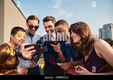 multiracial joyful people in casual clothes watching photos at smartphone at rooftop party Stock Photo