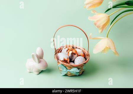 Basket with painted white and golden eggs, bunny and yellow tulips on light green background. Easter concept. Closeup. Stock Photo