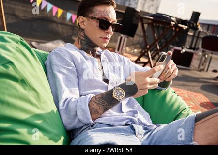 good looking young man with tattoos and sunglasses relaxing during party and looking at smartphone Stock Photo