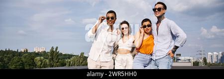 group of four joyous interracial friends with stylish sunglasses posing together on rooftop, banner Stock Photo