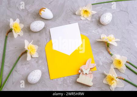 Yellow envelope with blank card, Easter eggs and daffodils flowers on textured gray background. Top view, flat lay, mockup Stock Photo