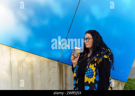 Beautiful young brunette woman with long curly hair, dressed in a black, blue and yellow waistcoat, drinks coffee from a paper cup on a background of Stock Photo