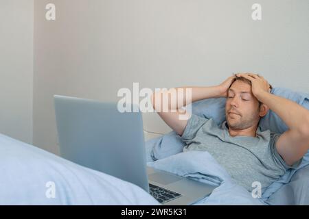 Shocked man working on laptop while lying in bed in the morning at home. Dumbfounded male freelancer hold his head with hands, have problems while Stock Photo