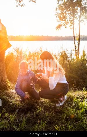 This heartwarming image captures a tender moment between a mother and her young child during a beautiful lakeside sunset. The soft, golden sunlight filters through the trees, illuminating the scene with a radiant glow. The mother, crouching down to her child's level, engages in a gentle and educational interaction, possibly exploring the nature around them. The child, dressed in a blue jacket, looks on with curiosity and wonder. The scenic backdrop of the calm lake and the surrounding foliage enhances the serene and nurturing atmosphere of the moment. Mother and Child Enjoying Lakeside Sunset  Stock Photo