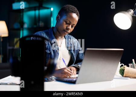 African American woman works diligently at her desk using her laptop and pen, researching, and brainstorming ideas. A productive and focused individual utilizing technology for work and study. Stock Photo