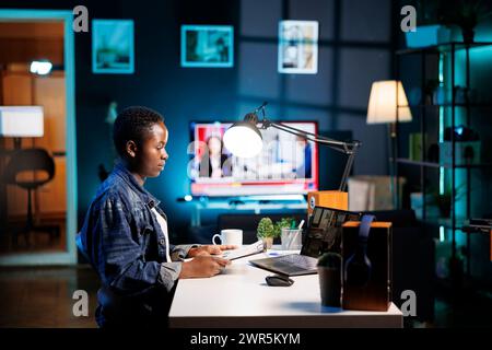 African american college student grasping paperwork while having a conversation through a laptop with female colleague. Two young women having an online discussion, video call on portable computer. Stock Photo