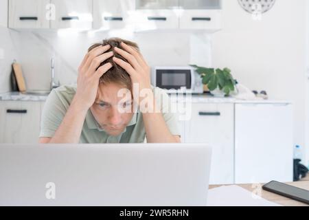 Disappointed exhausted man freelancer working at home on laptop, overworked, needs rest and break. Dumbfounded male freelancer hold his head with Stock Photo