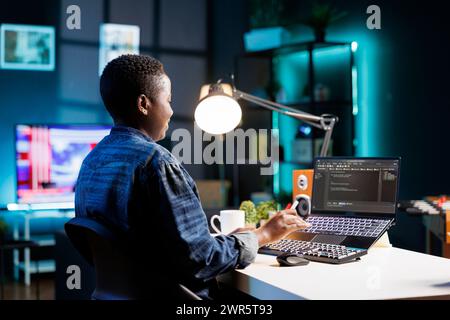 African American freelancer working at a desk focused on her laptop that is displaying code and data. Black woman with wireless computer, programs and manages the software, ensuring cyber security. Stock Photo
