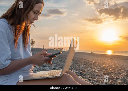 Digital nomad woman working with laptop and mobile phone on the beach by the sea during sunset. Female freelancer work online using smartphone and Stock Photo