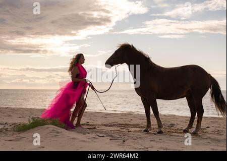 A woman in a pink dress standing beside a horse on the beach Stock Photo