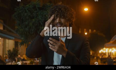 Young Indian Arabian Latino American man using smartphone mobile phone texting chatting online city street outdoors guy holding head problem headache Stock Photo