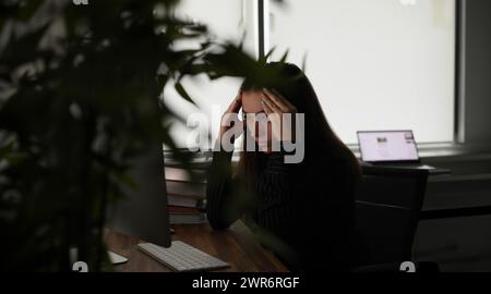 Tired woman holding her head with hands in front of computer screen in office Stock Photo
