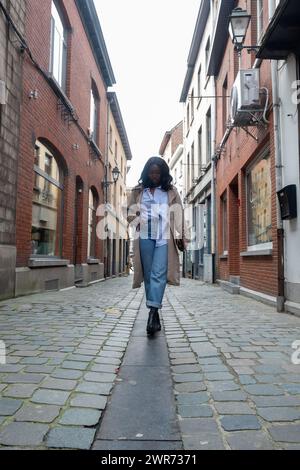 This image features a stylish woman walking down a cobblestone street in a historic European neighborhood. She is fashionably dressed in a blue collared shirt, high-waisted jeans, a beige trench coat, and black boots. Her confident stride and the perspective of the photograph suggest movement and a sense of purpose. The traditional buildings with brick facades and street lamps enhance the old-world feel of the setting. Stylish Woman Walking in Historic European Street. High quality photo Stock Photo