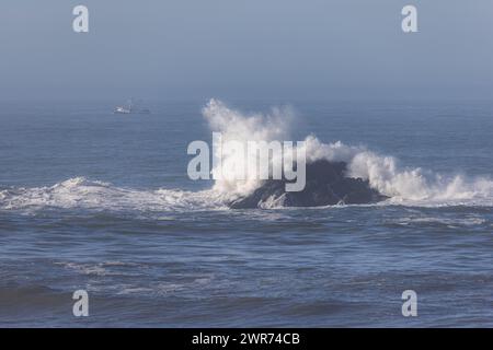 The ocean is calm and the water is blue. A large rock is in the water and the waves are crashing against it Stock Photo