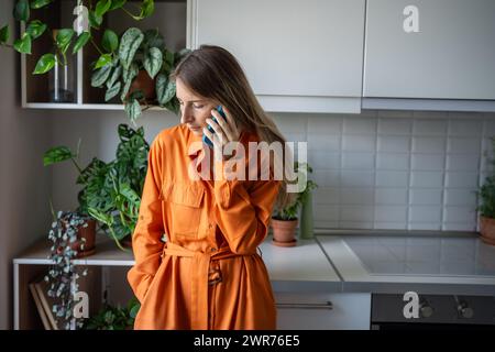 Concerned middle aged woman speaks on smartphone, sadly looking down while standing in home kitchen. Stock Photo