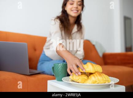 Smiling woman reaching for croissants on plate next to her laptop Stock Photo