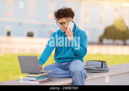 college student guy talks on phone browsing on laptop outdoors Stock Photo