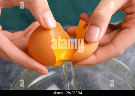 Housewife hands cracking fresh egg, yolk and white dropping in a bowl. A woman's hand cracking an egg with knife into a clear glass bowl in the gray k Stock Photo