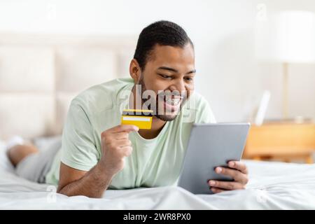 Black guy lying on bed, using digital tablet, bank card Stock Photo