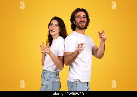 Laughing young woman and bearded man standing back to back, playfully pointing fingers at the camera Stock Photo