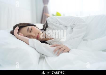 A beautiful woman lays her hands on a soft pillow. A young woman wearing white pajamas lies under a warm blanket. On a comfortable bed in the bedroom Stock Photo