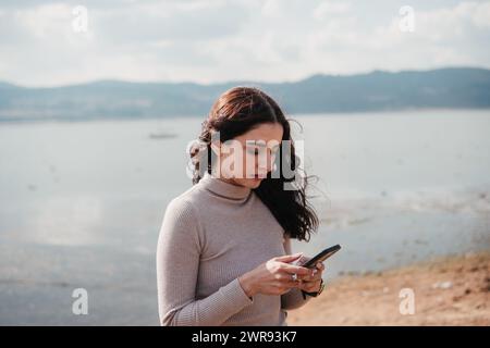 A focused young woman uses her smartphone near a calm lake, immersed in digital communication Stock Photo